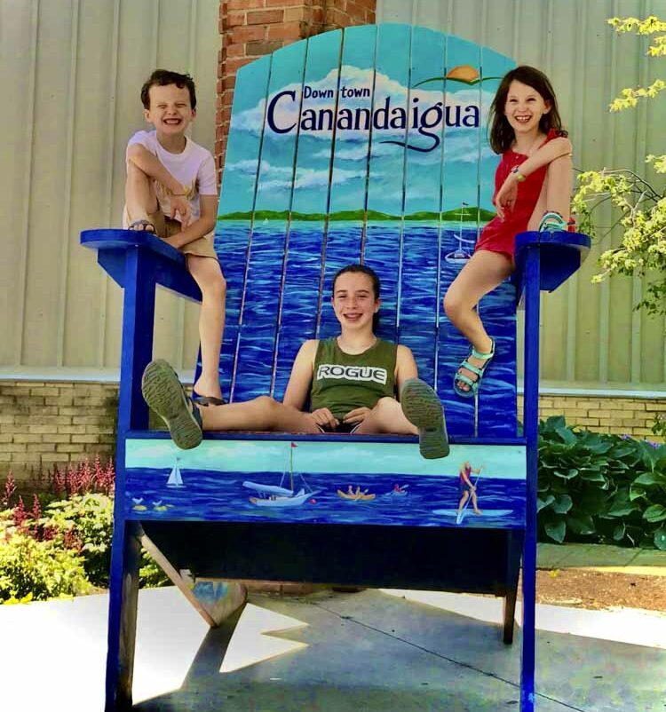 three children sitting on a large folding chair that reads Downtown Canandaigua